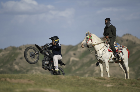 Man performing a wheelie on an electric bike while another person watches on horseback in a mountainous landscape, illustrating riding adventure and emphasizing Riding Times warranty protection.