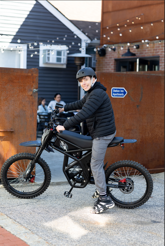 Young man in black helmet and jacket riding a black electric bike outdoors near apartments, showcasing e-bike riding times and speed benefits in 2025