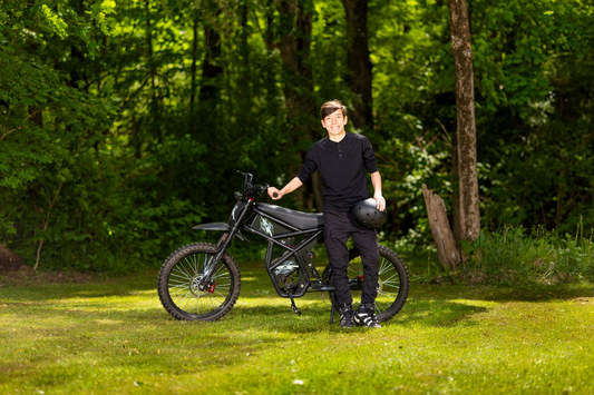 Teen rider standing next to black electric dirt bike in forest clearing promoting lithium battery safety for e-bikes
