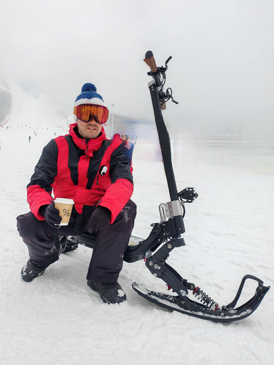 Man in winter gear sitting next to G63 Convertible Electric Scooter adapted with snow skis on snowy landscape
