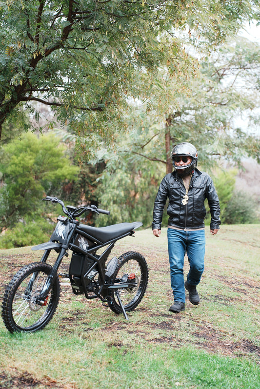 Man with helmet walking near a black electric dirt bike in a park setting for Riding Times eBikes Affiliate Program promotion