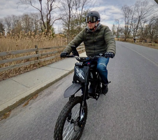 Man riding a black electric off-road motorcycle on a paved road wearing helmet and goggles