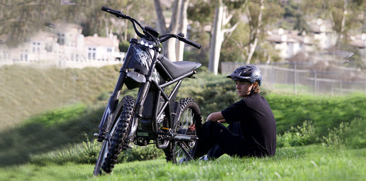 Young rider resting beside a rugged black electric dirt bike in a grassy park setting