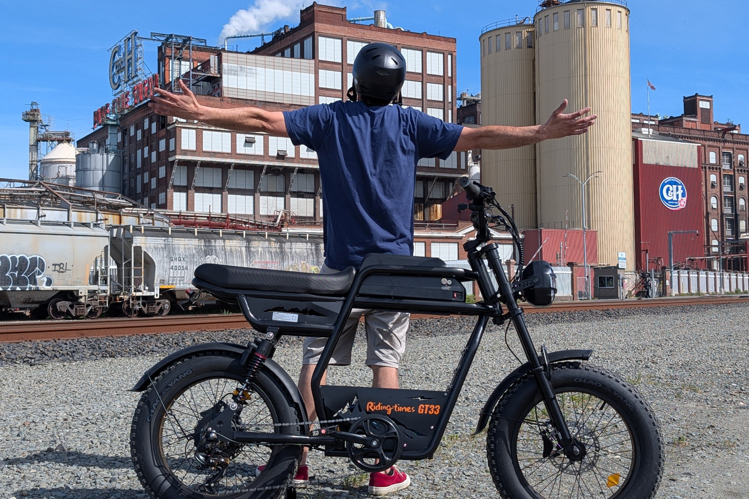 Rider with arms outstretched on the Riding Times GT33 all-terrain cargo electric bike near a railway yard, showing the bike's adventure-ready riding position