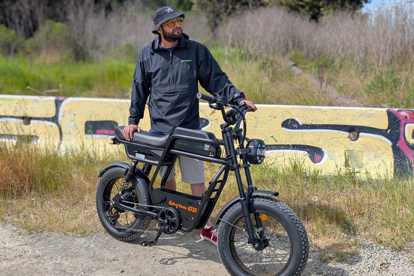 Rider standing beside the Riding Times GT33 cargo electric bike on a gravel path with a graffiti wall background, showing the front fork, fat tires, and open frame geometry