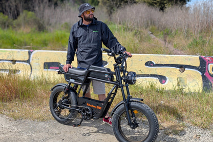 Rider standing beside the Riding Times GT33 cargo electric bike on a gravel path with a graffiti wall background, showing the front fork, fat tires, and open frame geometry