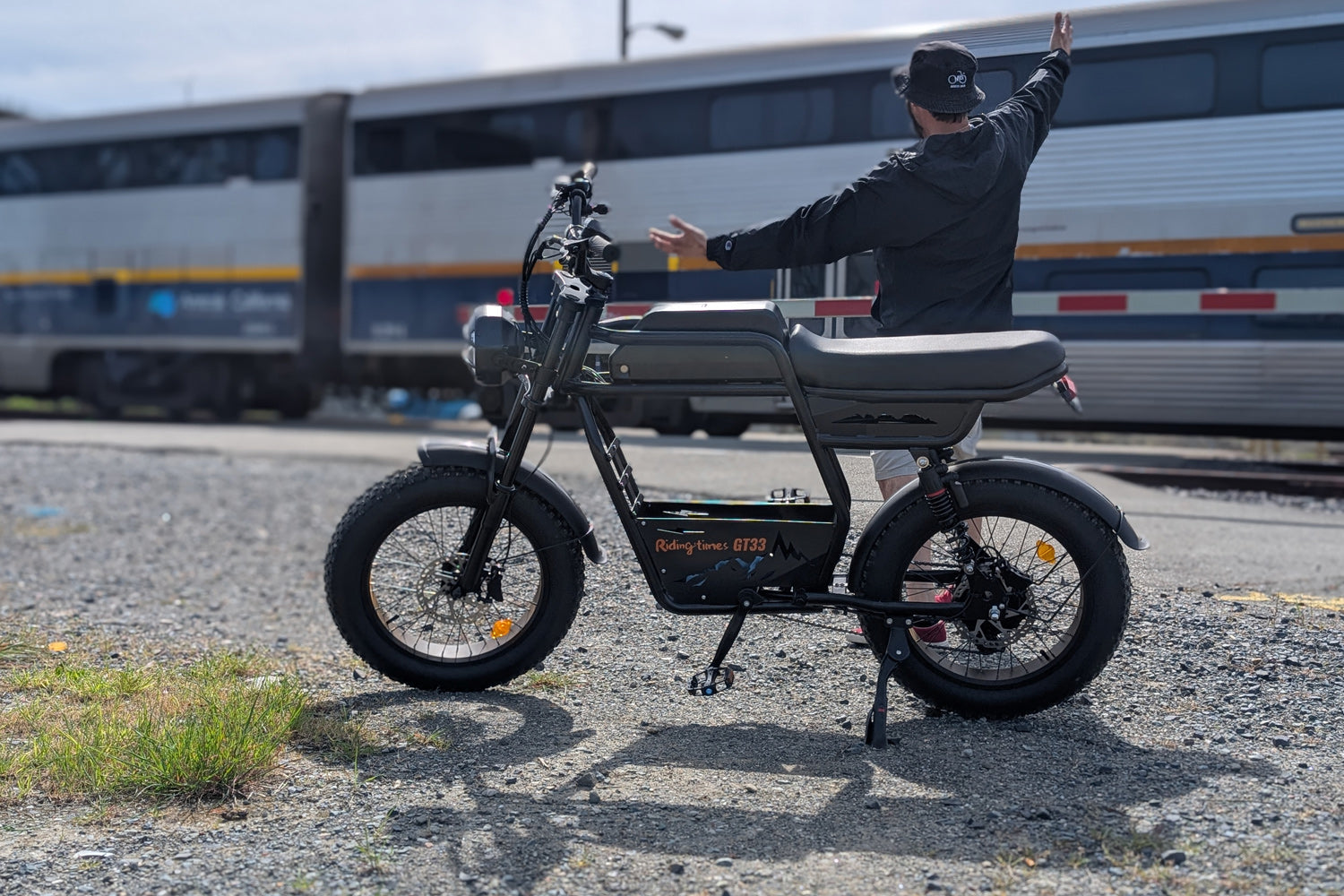 Rider seated on the Riding Times GT33 eBike near train tracks with an Amtrak train in the background, arms raised, highlighting the all-terrain fat tires and long flat seat
