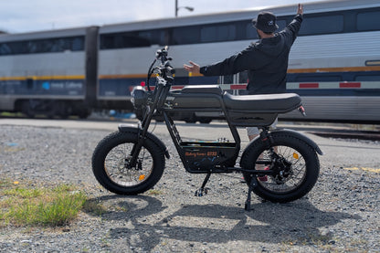 Rider seated on the Riding Times GT33 eBike near train tracks with an Amtrak train in the background, arms raised, highlighting the all-terrain fat tires and long flat seat