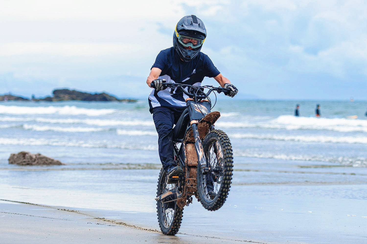 Person riding GT73 Pro electric motorbike on muddy wet beach performing a wheelie with ocean waves in background
